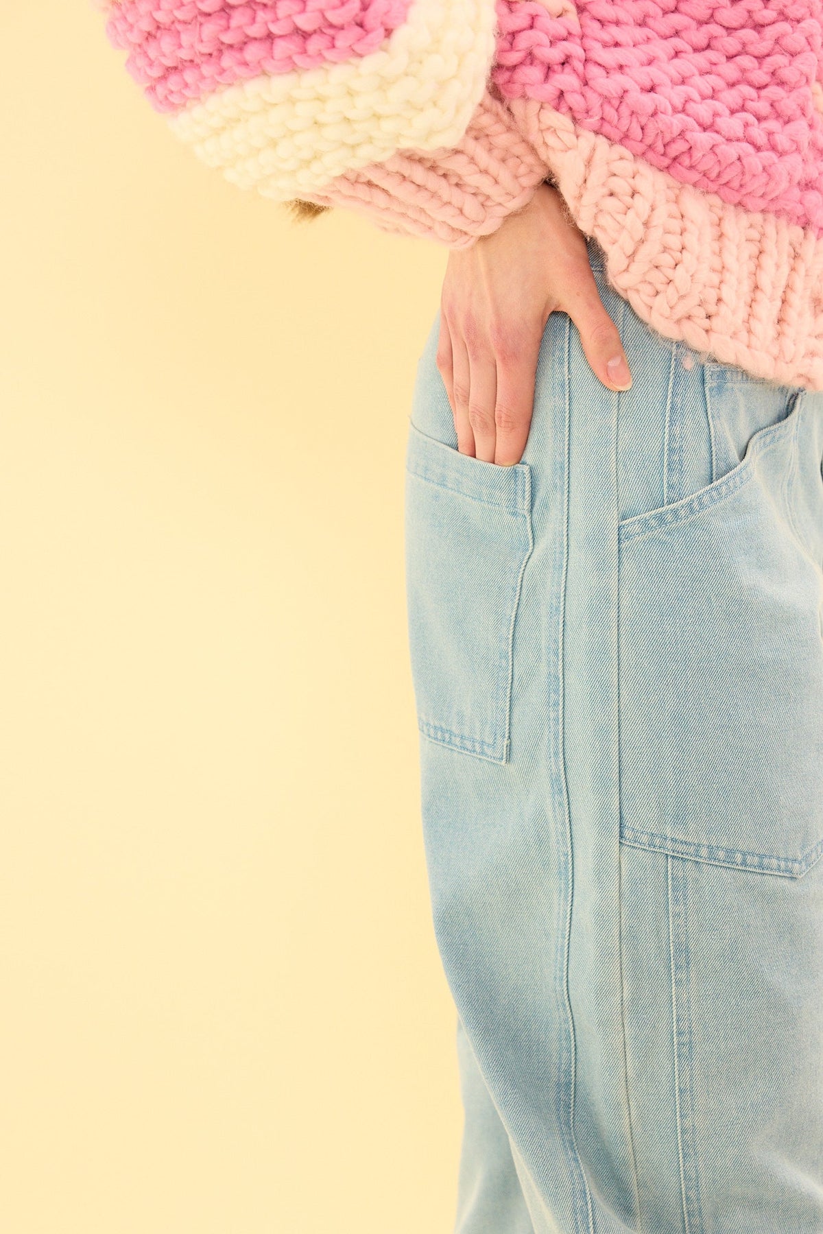 A person stands against a pale yellow background with one hand in their back pocket, wearing the Ivy City Co Amanda Pants in Light Wash Denim and a chunky pink sweater.
