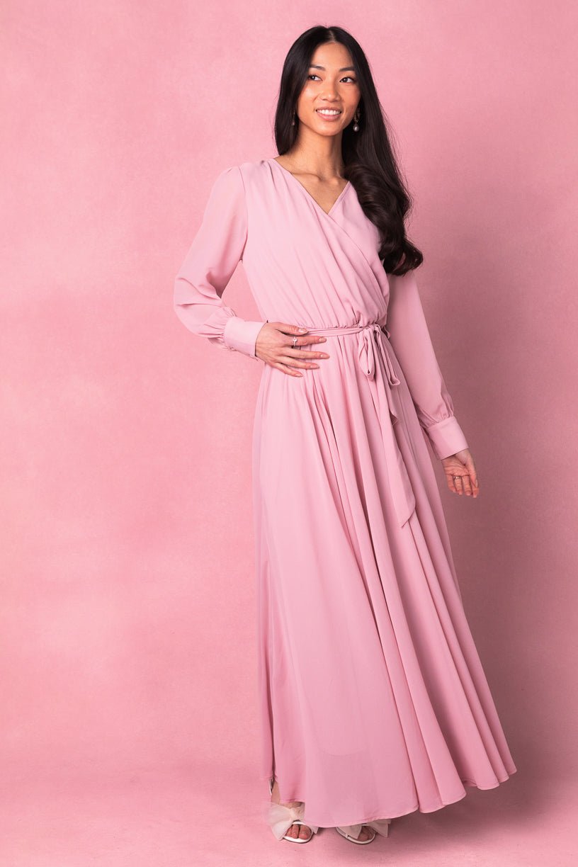 A woman with long dark hair smiles against a pink background, wearing the Ivy City Co Andie Dress in Blush—a maxi dress featuring long sleeves and a tied waist.