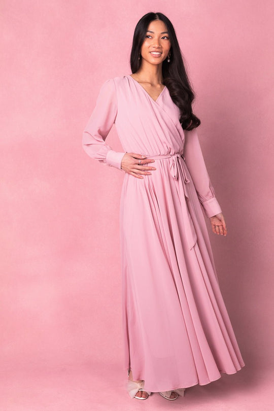 A woman with long dark hair smiles against a pink background, wearing the Ivy City Co Andie Dress in Blush—a maxi dress featuring long sleeves and a tied waist.