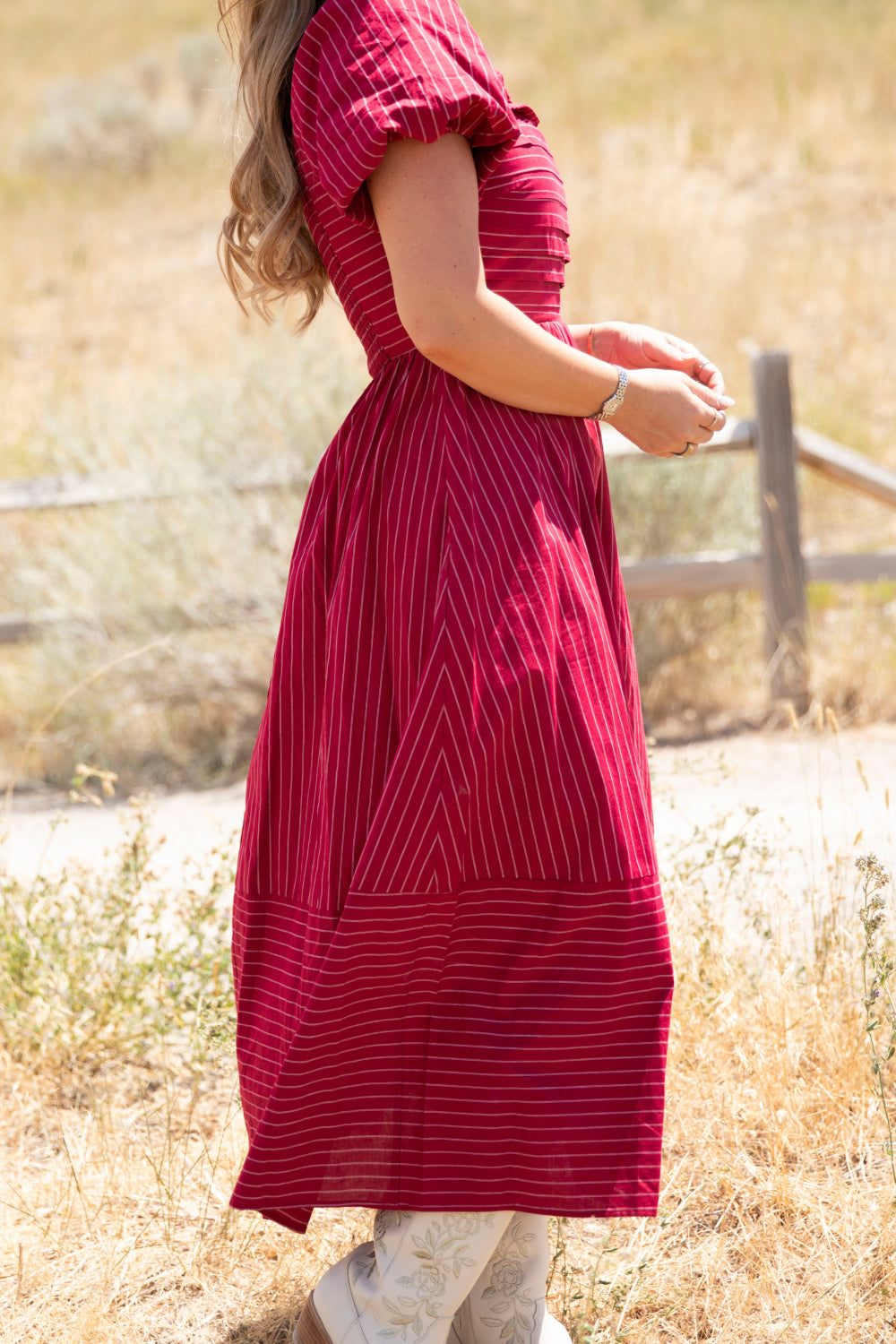 A woman stands outdoors on a sunny day, hands together and facing sideways, wearing the Ivy City Co Julie Dress in Cranberry Pinstripe. The dress features a smocked bodice and puff sleeves, adding charm to her look. A wooden fence and dry grass are visible in the background.