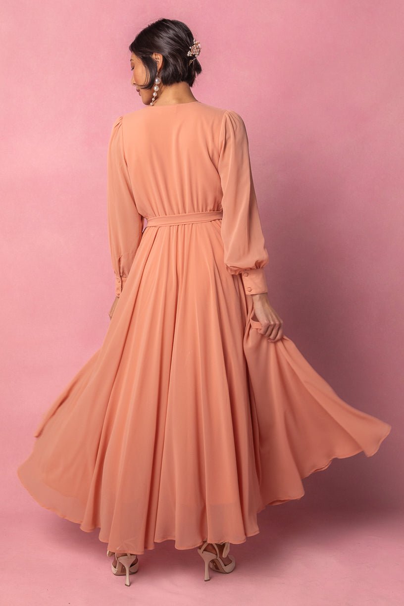 A woman stands against a pink background, facing away as she holds the skirt of the Ivy City Co Andie Dress in Apricot Crush with one hand. She wears heels and has her hair styled up with a decorative hairpiece.