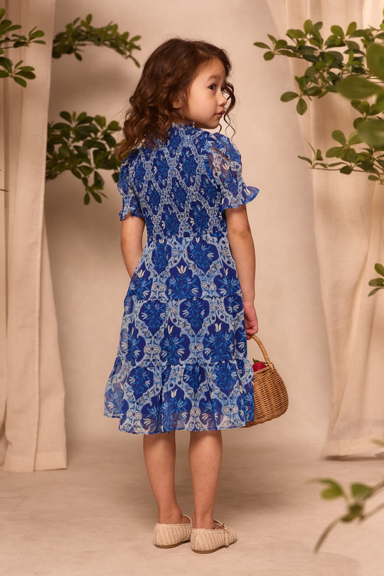 A young girl with curly hair stands indoors among lush green plants and beige curtains, holding a small wicker basket. She wears the Ivy City Co Mini Cordelia Dress in Trellis Foulard—a stunning blue floral piece perfect for any occasion. This elegant girls chiffon dress is an ideal choice for matching family outfits or anyone seeking elevated, charming style.