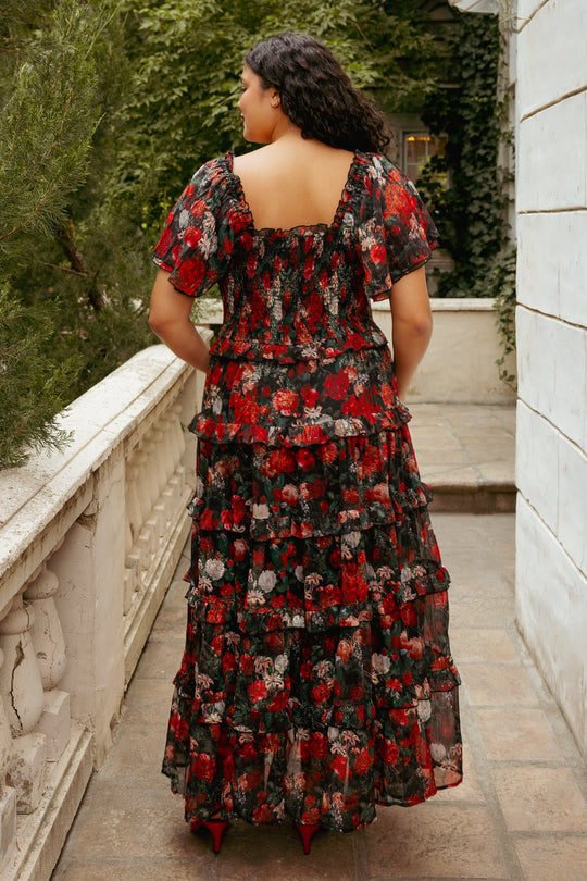 A woman with long, dark hair stands outdoors, facing away. She wears the flowy, tiered Ivy City Co Winnie Dress in Midnight Roses—featuring a striking black base adorned with a vibrant midnight roses pattern and short sleeves. This maternity-friendly dress stands out beautifully against the green foliage and white wall in the background.