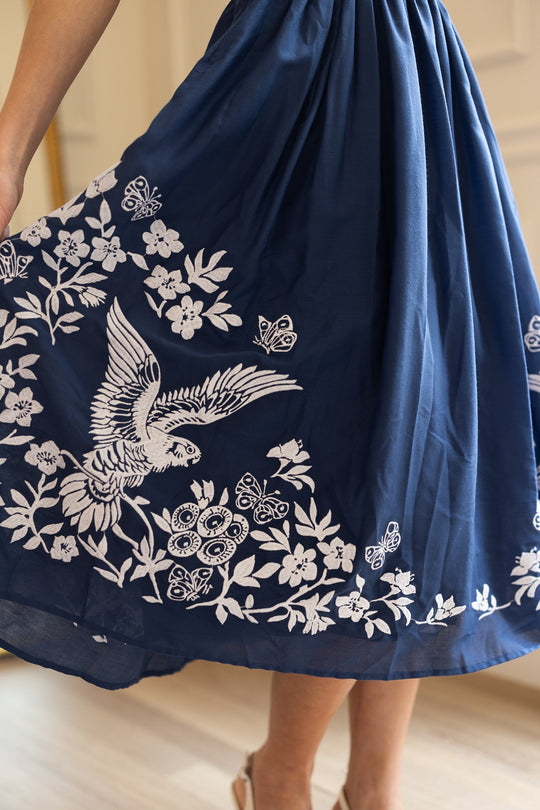 A person wearing the Ivy City Co Finch Dress in Blue, featuring white embroidery with designs of an owl, a bird in flight, flowers, and butterflies. Only the lower half of the skirt and legs are visible.
