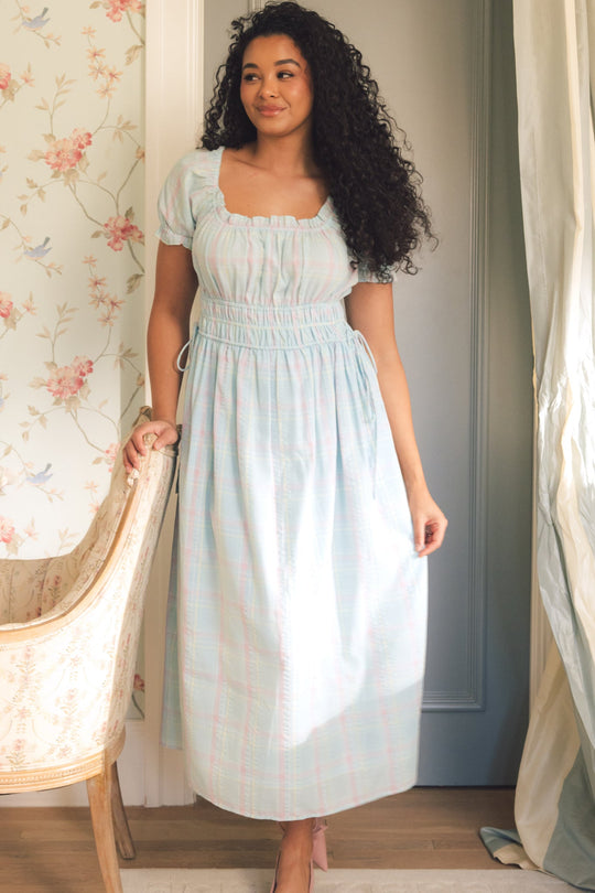 A woman with long, curly hair stands indoors by a floral armchair, smiling softly as she wears the Thalia Dress in Pastel Plaid from Ivy City Co, surrounded by floral wallpaper and matching curtains.