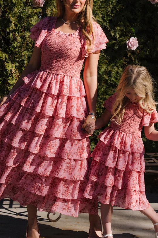 A woman and a young girl hold hands, both wearing matching Ivy City Co Grace Dress in Desert Heritage Rose. These flutter-sleeve maxi dresses feature ruffled details and a pink floral print, making them perfect for outdoor strolls among greenery and flowers. The design is also maternity friendly.
