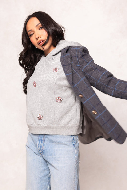 A woman with long dark hair wears the Ivy City Co Skylar Hoodie in Heather Gray, featuring crystal flower embellishments, paired with light blue jeans and a blue plaid blazer draped over one shoulder as she poses against a light background.