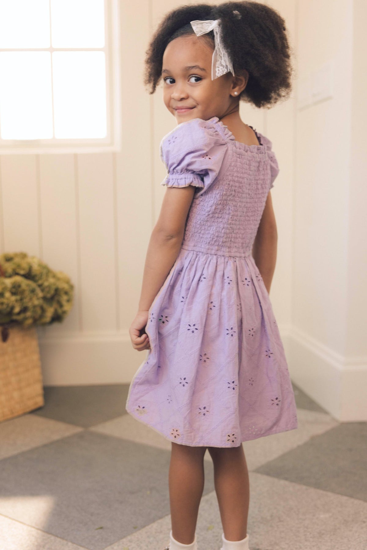 A young girl with curly hair and a white bow wears the Ivy City Co Mini Emmi Dress in Lilac, featuring delicate eyelet details. She stands indoors on patterned tile flooring, smiling over her shoulder, while a basket of dried flowers in the background completes her charming springtime look.