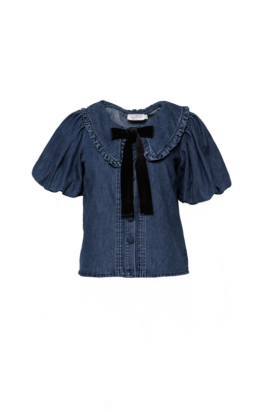 The Andrea Blouse in Denim with Velvet Tie by Ivy City Co features puffed short sleeves, a ruffled collar, front buttons for nursing-friendly wear, and a black velvet neck bow, shown on a white background.