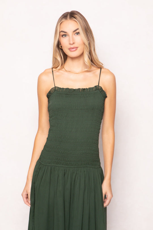 A woman with long blonde hair is wearing the Ivy City Co Kimberly Dress in Green—a dark green midi dress with a maternity-friendly, smocked bodice and spaghetti straps—standing against a light background and looking at the camera with a neutral expression.