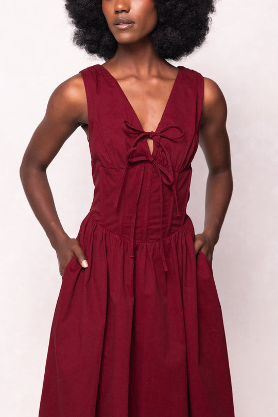 A woman stands with her hands in the pockets of the Ivy City Co Kayla Dress in Burgundy—a sleeveless dress featuring a deep red hue, v-neckline, and a tie detail at the chest—posed against a light background.