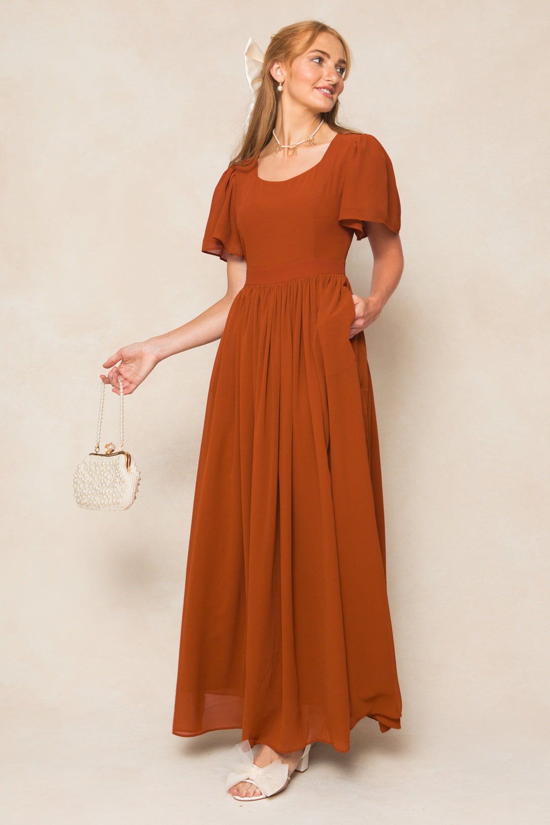 A woman in the Ivy City Co Abbie Dress in Amber Chiffon holds a small cream handbag, smiles, and stands against a neutral background. She accessorizes with white sandals and a white bow in her blonde hair.