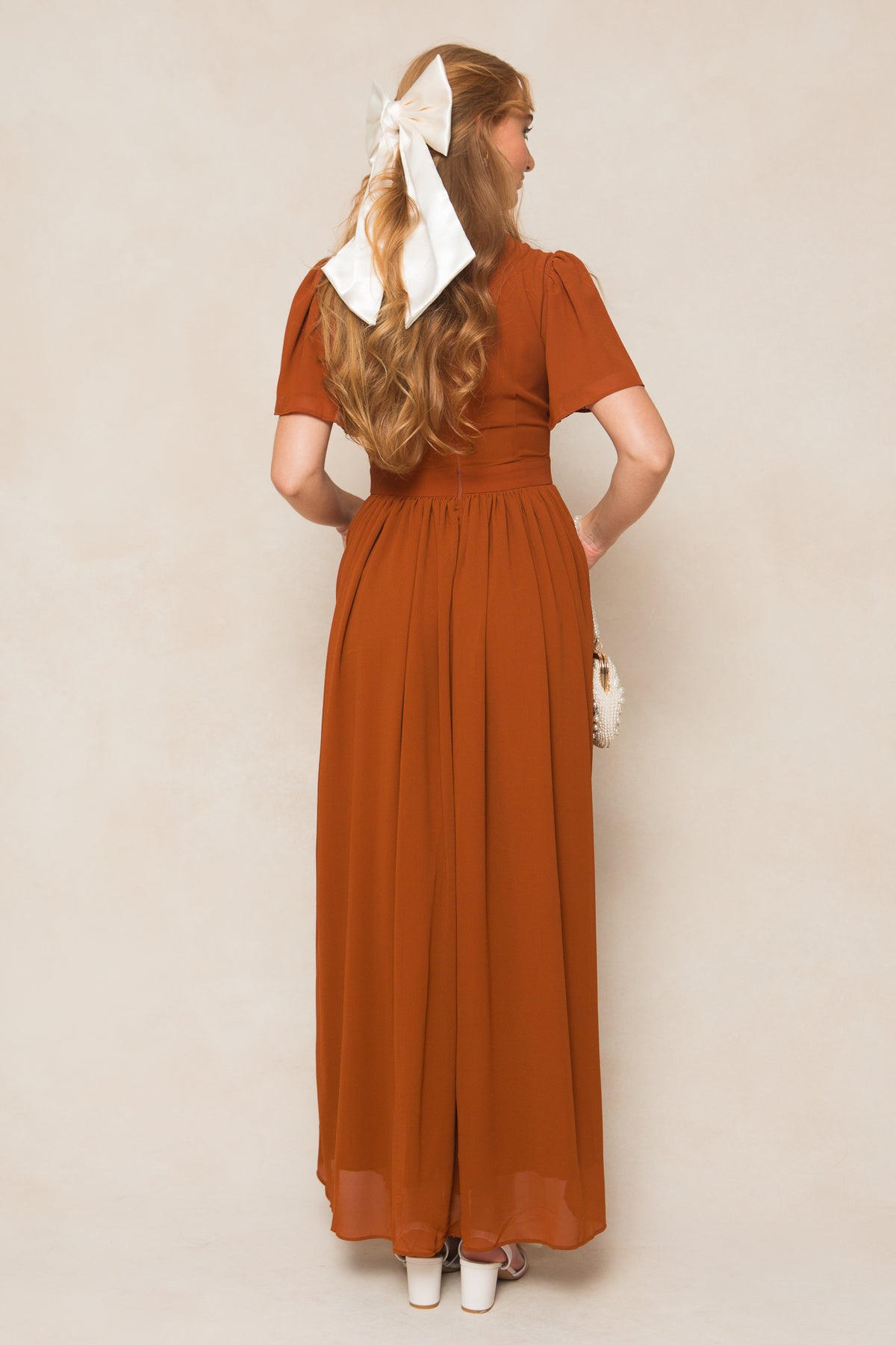 A woman with long hair and a white bow stands with her back to the camera in the Ivy City Co Abbie Dress in Amber Chiffon, a short-sleeve maxi dress, paired with a small white purse and white platform heels.