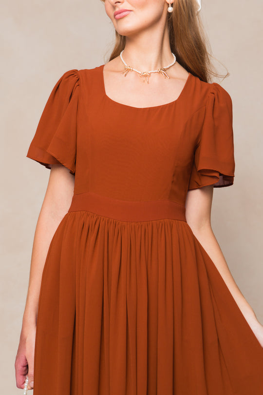 A woman wears the Ivy City Co Abbie Dress in Amber Chiffon, featuring short sleeves, a square neckline, and a gathered skirt. She accessorizes with pearl jewelry; her face is partially out of frame against a neutral background.