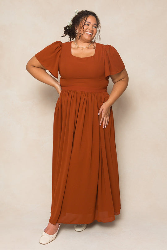 A woman stands smiling against a beige background, wearing the Ivy City Co Abbie Dress in Amber Chiffon—a short-sleeved, maxi-length dress with a square neckline. Her curly hair is partially tied back, and she completes the look with cream-colored flat shoes.