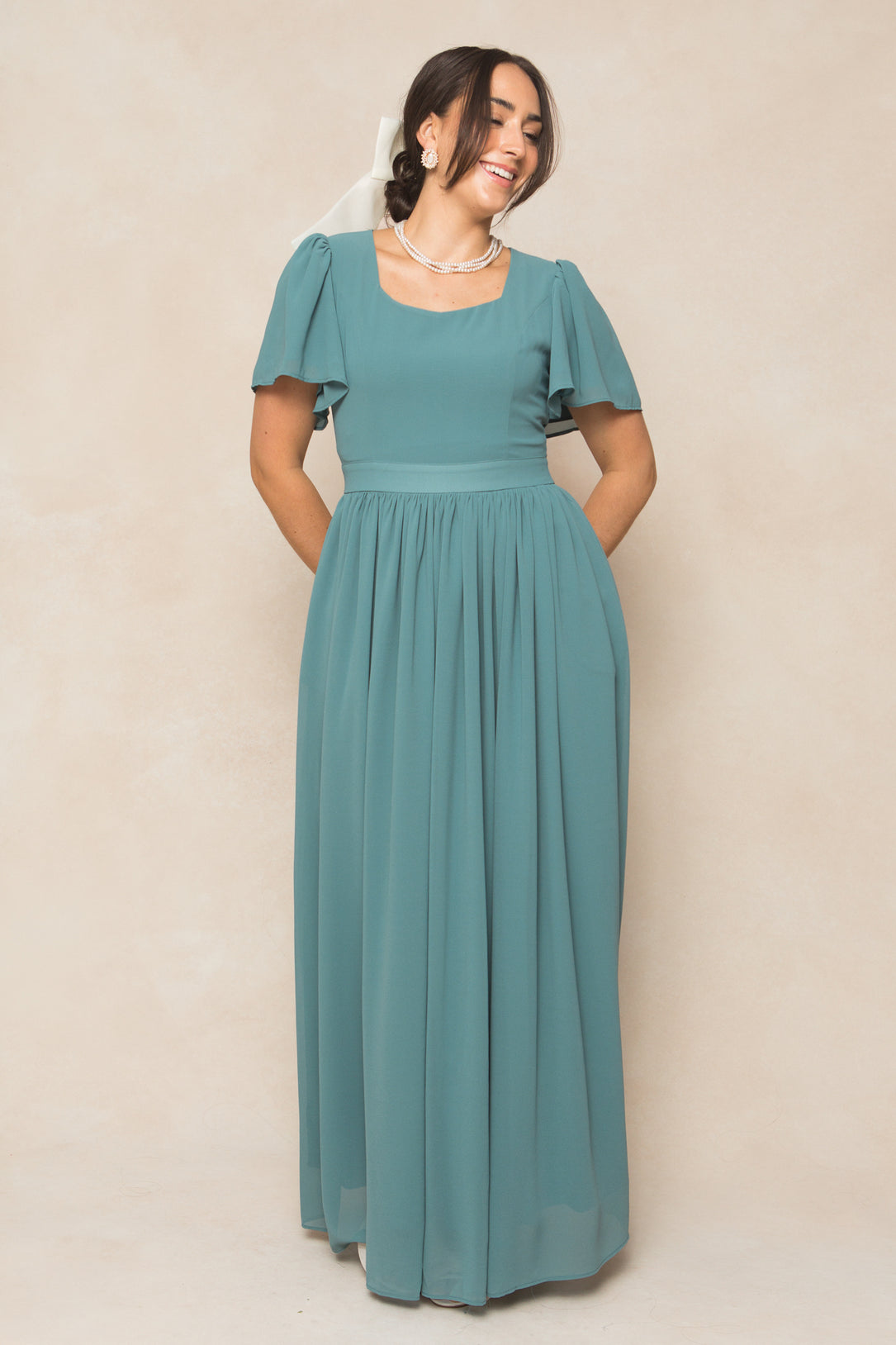 A woman smiles in the Ivy City Co Abbie Dress in Seaglass Chiffon—a floor-length gown with flutter sleeves, a fitted waist, and square neckline. She accessorizes with pearls and a white bow against a neutral backdrop. Ideal for fall bridesmaids.