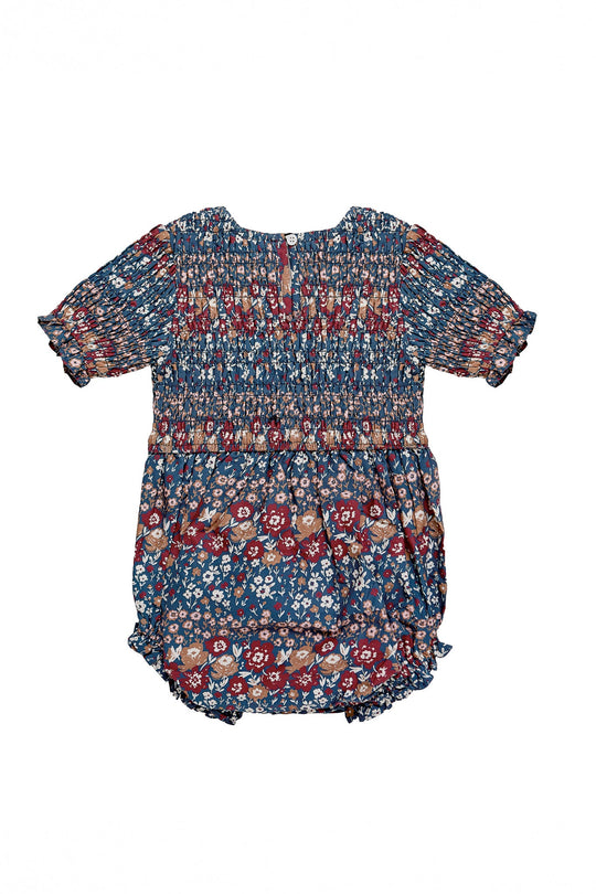 The Ivy City Co Baby Parker Romper in Dusk Floral Medley features short sleeves, ruffled leg openings, and an elegant smocked bodice. Made from lightweight cotton, this romper displays a blue base covered in a delicate multicolored floral print in shades of red, white, and brown. The garment is shown laid flat on a white background to clearly highlight its details.