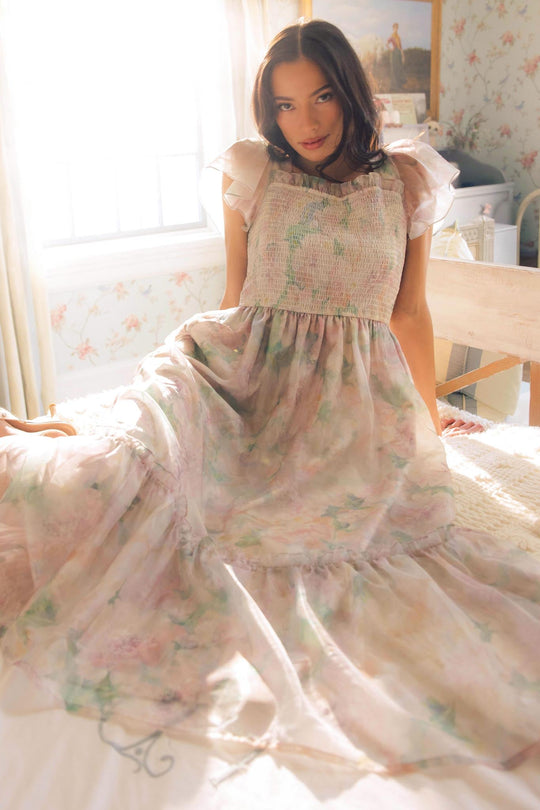 A woman wearing the Ivy City Co Rhiannon Dress in Painted Bloom sits on a bed in a sunlit, vintage-style room with floral wallpaper, looking at the camera.