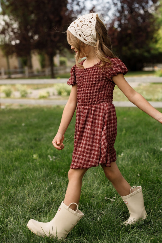 A young girl walks on grass outdoors, holding a small twig, wearing the Ivy City Co Mini Tori Dress in Cranberry Popcorn Plaid with puff sleeves, paired with cream boots and a white knit headscarf. Trees and a blurred background are visible behind her.