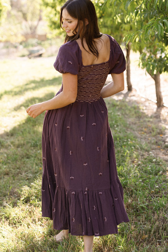 A woman in the Ivy City Co Acantha Dress in Aubergine Embroidered Gauze, featuring puff sleeves and a maternity-friendly fit, stands barefoot on grass, turning to the side with her back partially facing the camera, outdoors among sunlit trees.