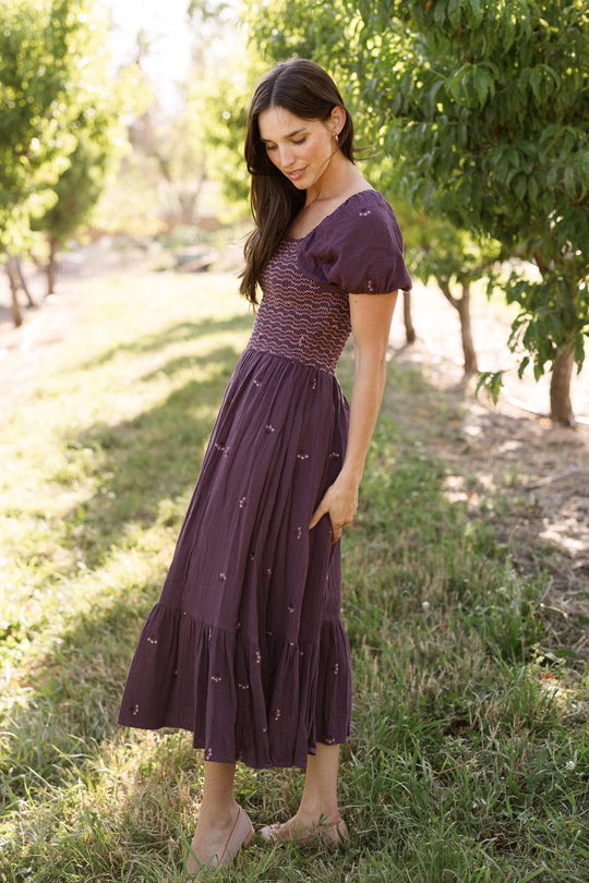 Wearing the Ivy City Co Acantha Dress in Aubergine Embroidered Gauze—a purple, short-sleeved style with subtle puff sleeves and a smocked bodice—a woman stands on grass in a sunlit orchard, looking down as she gently touches her skirt. This elegant dress is also maternity friendly, making it an ideal choice for warm days outdoors.