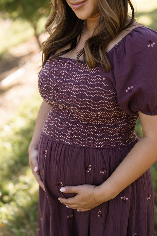 An expectant woman stands outdoors in an Ivy City Co Acantha Dress in Aubergine Embroidered Gauze, gently cradling her belly with both hands. Her long, wavy hair frames her partially visible face, while the blurred green background draws attention to the dress’s comfortable, maternity-friendly design and elegant puff sleeves.