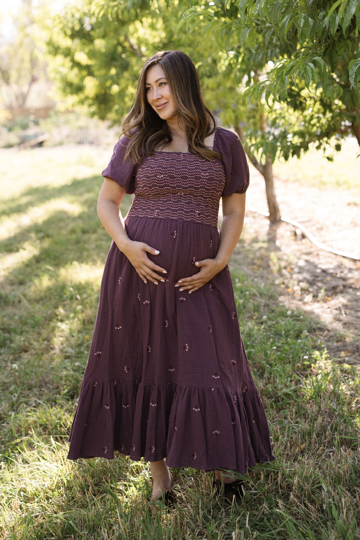 A woman in the Ivy City Co Acantha Dress in Aubergine Embroidered Gauze with puff sleeves stands barefoot on grass, cradling her belly with both hands. She smiles softly and gazes to the side as sunlight filters through the trees in the background.