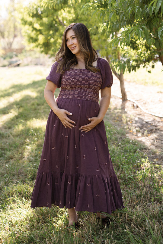 A woman in the Ivy City Co Acantha Dress in Aubergine Embroidered Gauze with puff sleeves stands barefoot on grass, cradling her belly with both hands. She smiles softly and gazes to the side as sunlight filters through the trees in the background.