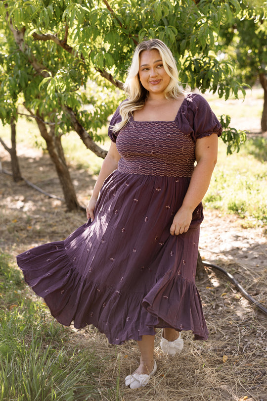 A woman with long blonde hair stands outdoors in a sunny, green area, smiling and holding the edge of her dress. She is wearing the Ivy City Co Acantha Dress in Aubergine Embroidered Gauze, which features puff sleeves and an elegant, maternity-friendly design.