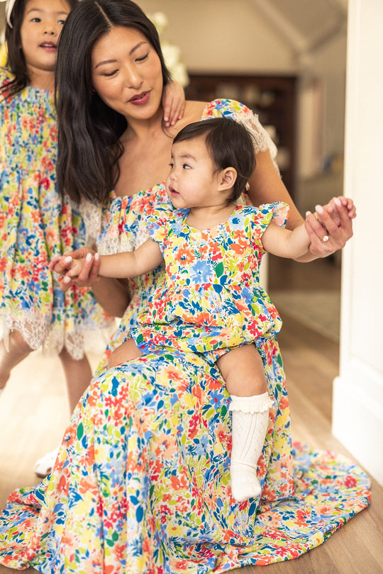 A woman and two young children wear matching Ivy City Co Ada Dresses in Vibrant Yarrow Print. The woman kneels, smiling at the baby in her lap, while the older child stands nearby. Their coordinated Ada Dresses are both nursing friendly and maternity friendly, making them perfect for family moments together indoors on a wooden floor.