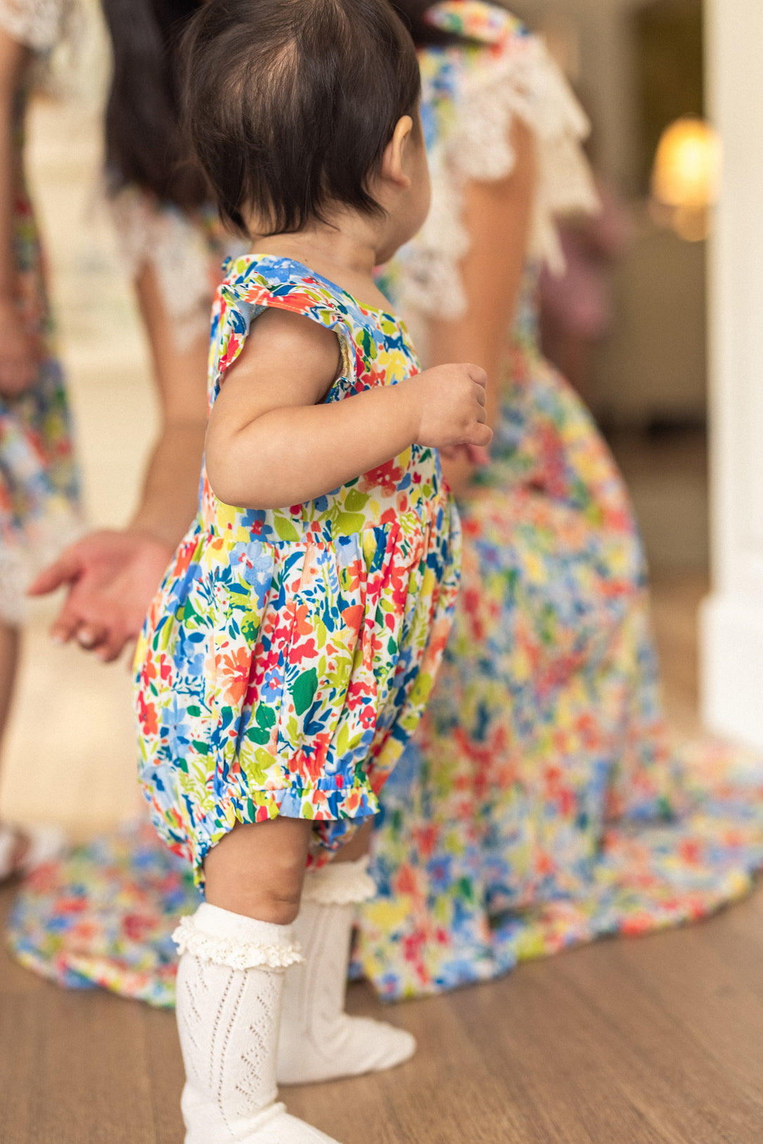 A baby in the Ivy City Co Baby Ada Romper in Vibrant Yarrow Print and white knee-high socks stands on a wooden floor, facing away, with adults in matching floral looks visible in the background.