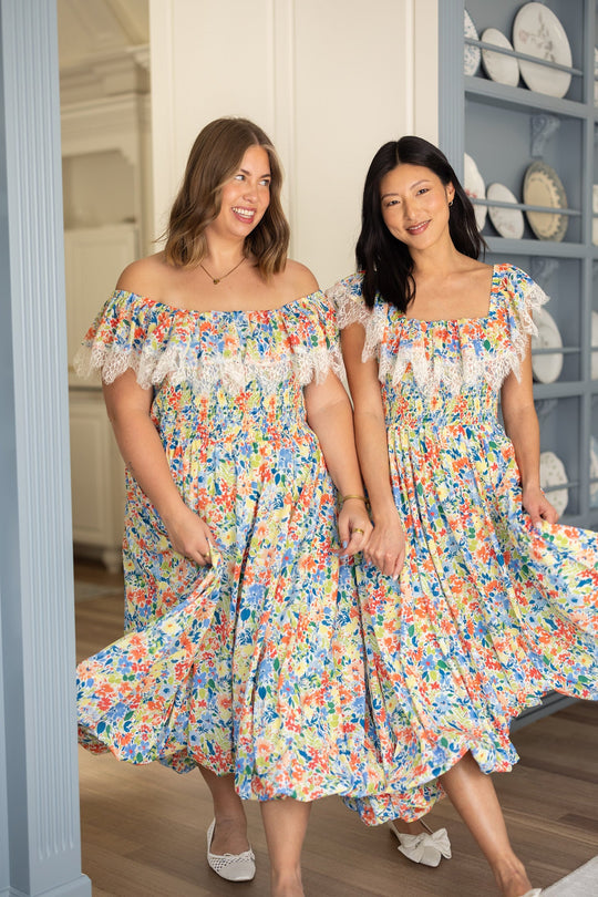 Two women holding hands, wearing matching Ivy City Co Ada Dresses in Vibrant Yarrow Print—a maternity-friendly, off-the-shoulder floral chiffon style with lace trim—smile as they walk indoors on wooden floors near blue shelves displaying white plates. These dresses offer both comfort and style for a joyful stroll.