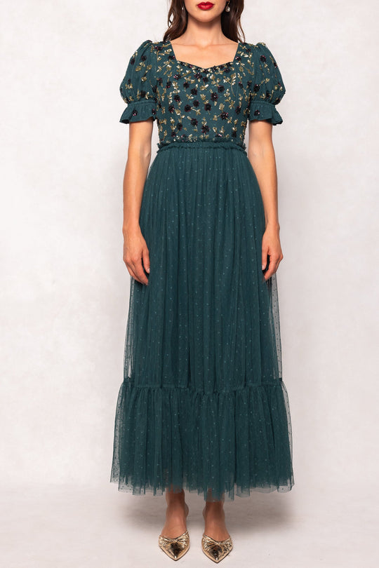 A woman wearing the Ivy City Co Adara Dress in Green—a teal, ankle-length tulle gown featuring floral embroidery on the bodice and puffed short sleeves—paired with gold pointed shoes, stands against a plain light backdrop.