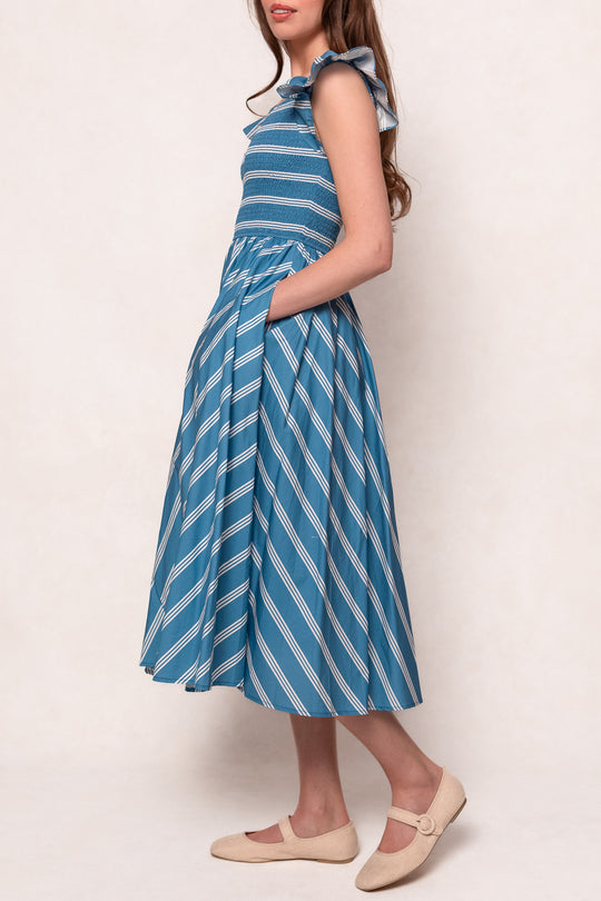 A woman stands sideways wearing the Ivy City Co Adelaide Dress in Blue, a striped cotton midi with ruffled shoulders and pockets, paired with beige flats.