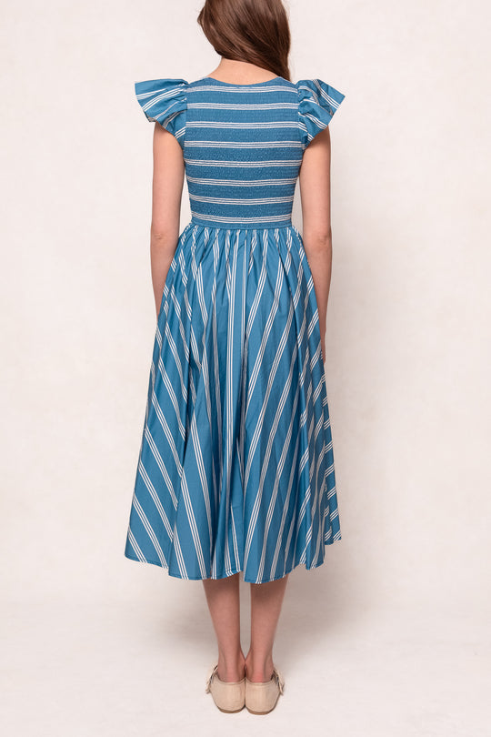 A woman stands facing away in the Ivy City Co Adelaide Dress in Blue, a cotton midi with flutter sleeves, pleated skirt, horizontal stripes on the bodice, convenient pockets, and light-colored flats.