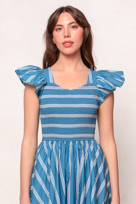 A woman stands facing forward against a plain light background, wearing the Ivy City Co Adelaide Dress in Blue—a cotton midi dress with blue and white stripes and ruffled sleeves.