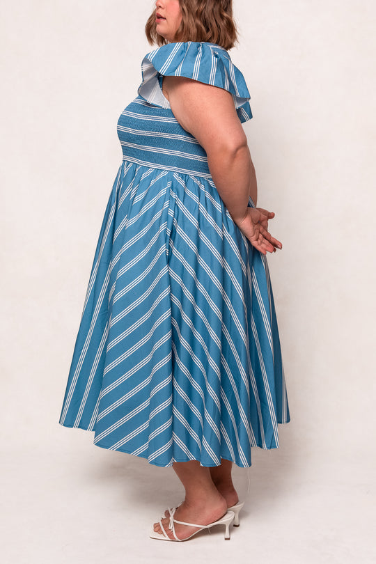 A woman models the Ivy City Co Adelaide Dress in Blue, featuring blue and white stripes and pockets. She stands sideways with hands behind her back, pairing the look with white open-toe heels against a light background.