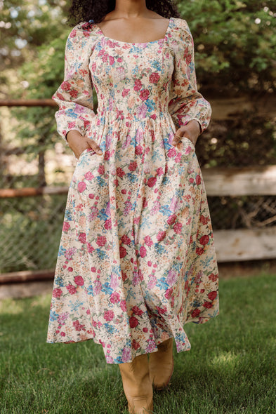 A woman stands on grass with her hands in the pockets of the Adelpha Dress in Autumn Amity Floral by Ivy City Co—a vintage-inspired, long-sleeve cotton dress featuring a fitted bodice and timeless floral print. The dress is maternity and nursing friendly, combining elegance with comfort. In the background are a wire fence and lush greenery.