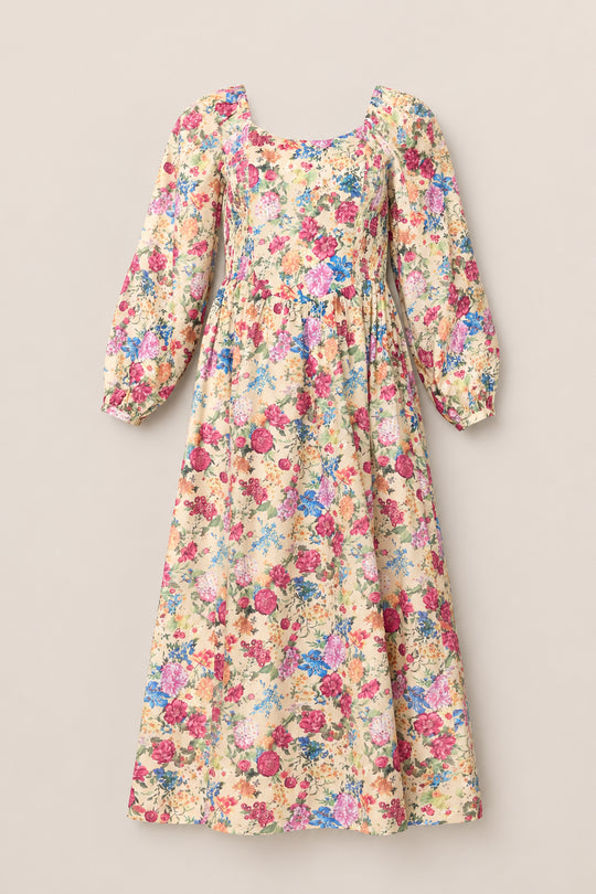 The Adelpha Dress in Autumn Amity Floral by Ivy City Co is a long, cream-colored maternity and nursing-friendly dress crafted from soft cotton, featuring a colorful floral print, puffed long sleeves, a square neckline, and a gathered waist. Displayed against a plain beige background.
