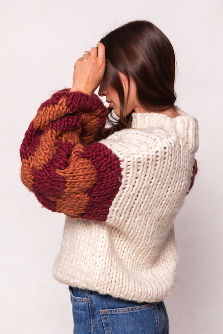 A woman in the Ivy City Co Aimee Sweater in Orange & Cream—an oversized wool blend with thick, textured sleeves—stands in profile, touching her head. She styles it with blue jeans and lets her long brown hair fall against a light background.