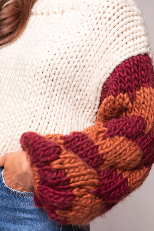 A close-up of a person wearing the Aimee Sweater in Orange & Cream by Ivy City Co—a chunky wool blend with thick cream body and textured orange sleeves—stylishly paired with blue jeans.