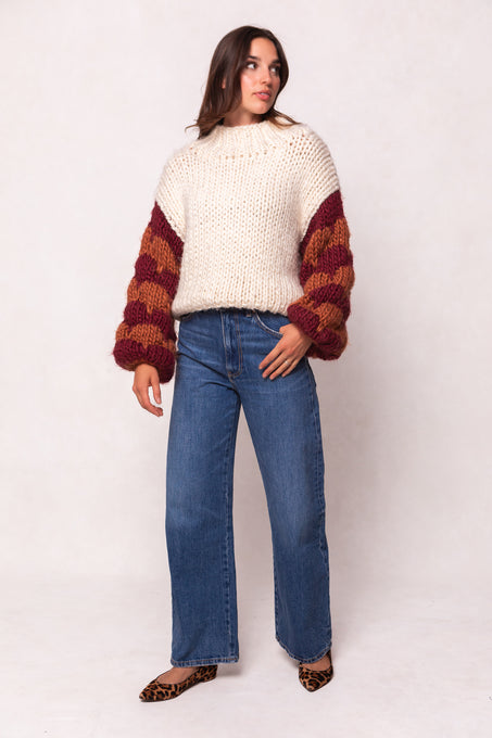 A woman with brown hair stands against a light background, wearing the Ivy City Co Aimee Sweater in Orange & Cream with maroon and orange sleeves, paired with blue wide-leg jeans and leopard print flats, looking slightly to the side.