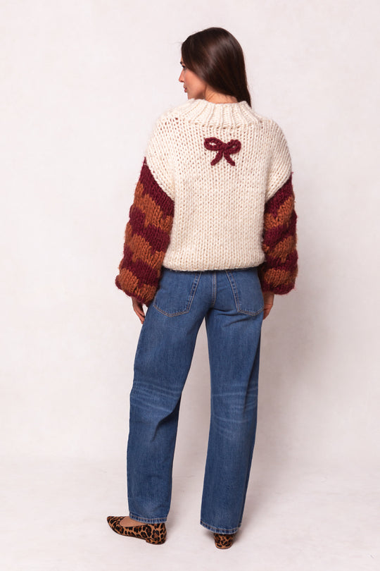 A woman faces away, wearing the Ivy City Co Aimee Sweater in Orange & Cream with oversized fit and maroon bow detail on back, paired with blue jeans and leopard print flats.