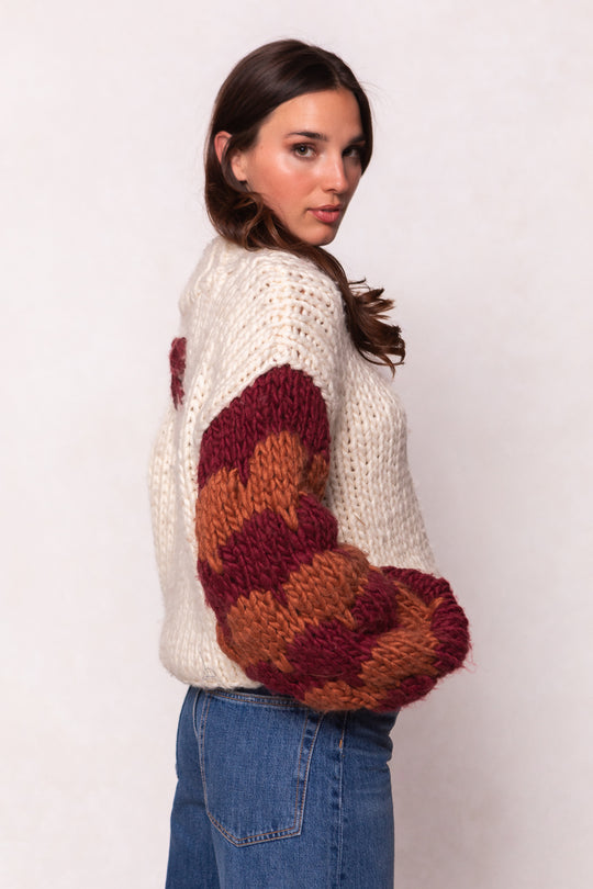 A woman with long brown hair wears the Ivy City Co Aimee Sweater in Orange & Cream, a chunky knit wool blend with bold striped sleeves, paired with blue jeans. She stands sideways, looking over her shoulder.