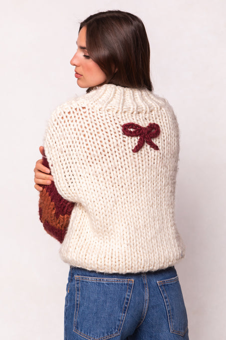 A woman with long brown hair wears Ivy City Co’s Aimee Sweater in Orange & Cream—an oversized wool blend with a small maroon bow on the upper back—paired with blue jeans and turned sideways to showcase the detail.