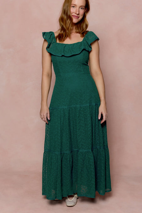 A woman with long, wavy hair smiles gently against a pale pink background, wearing the Ivy City Co Alaina Dress in Deep Sea Lace—an ankle-length dress featuring a ruffled neckline and tiered skirt, with one hand by her side.