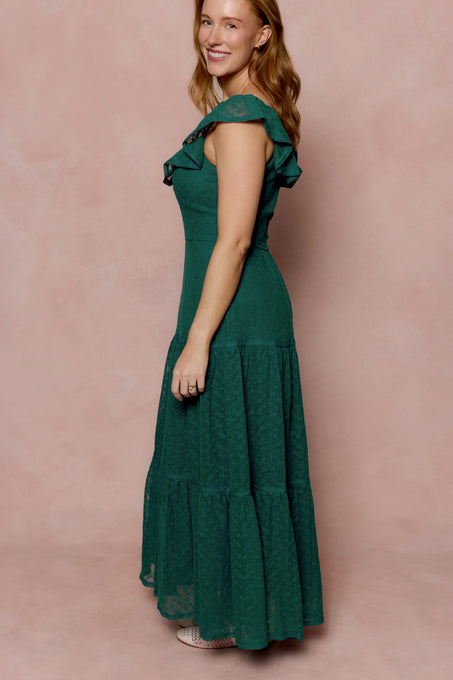 A woman with long, light brown hair smiles sideways in the Ivy City Co Alaina Dress in Deep Sea Lace—a floor-length gown with cap sleeves and ruffle details—posing against a soft pink background.