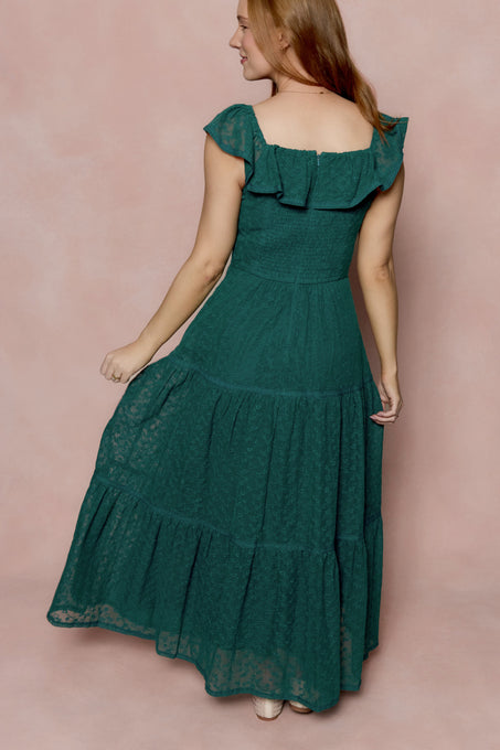 A woman with light brown hair wears the Ivy City Co Alaina Dress in Deep Sea Lace—an ankle-length, green, ruffled, cap-sleeve dress—as she stands sideways and looks over her shoulder against a soft pink background.