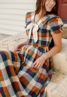 A woman sits on stone steps, smiling and looking down, wearing the Ivy City Co Alice Dress in Maize Gingham with puff sleeves and a large white bow at the collar.