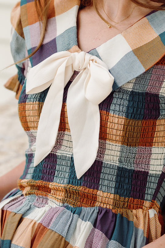 A person wearing the Ivy City Co Alice Dress in Maize Gingham, which features a smocked bodice, ruched plaid fabric, puff sleeves, and a large white bow at the collar.